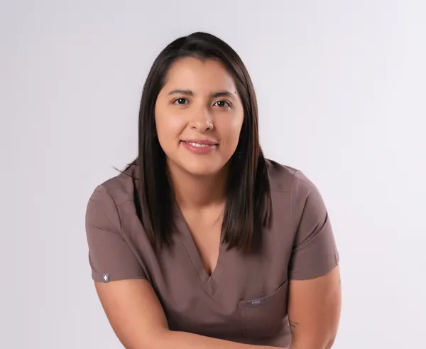 La Dra. Lourdes Alanis Naranjo, sentada con uniforme quirúrgico café, se inclina hacia la cámara y sonríe.