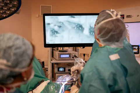 Cirujanos con gorro y cubrebocas operan por laparoscopía mientras siguen la cirugía en el monitor.