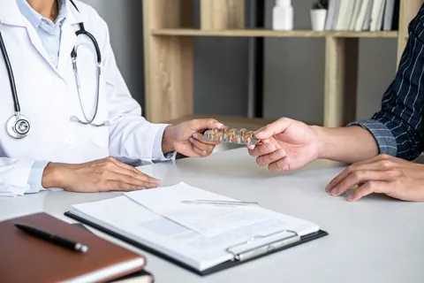 En el escritorio del consultorio, una mano con bata médica entrega un medicamento a otra persona junto a una receta.
